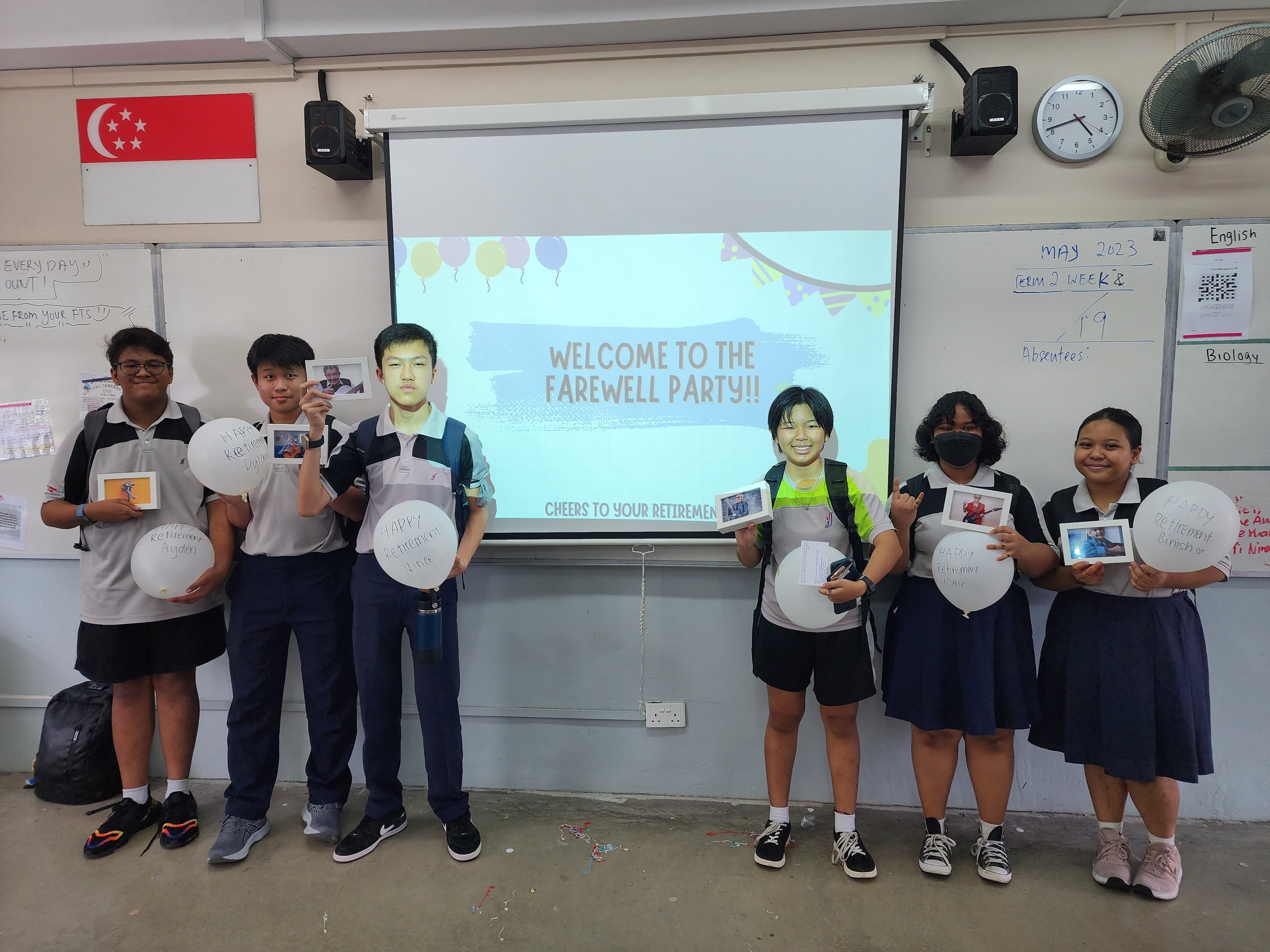 Six students in uniforms hold balloons and photos in a classroom with a projected farewell party slide behind them.